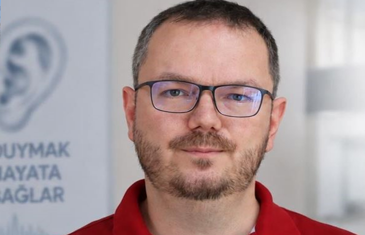 Görselin üst bölümünde, kırmızı polo yaka tişört giymiş, dikdörtgen koyu çerçeveli gözlük takan ve kirli sakallı bir erkek uzman hekimin doğrudan kameraya baktığı portresi yer alıyor. Arka planda aydınlık bir klinik veya ofis ortamı ile sol tarafta kulak çizimi ve "DUYMAK HAYATA BAĞLAR" sloganının bir kısmı okunan bilgilendirici dikey bir afiş görülüyor.