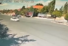 Görselin üst bölümünde, gündüz saatlerinde geniş bir caddeyi gören güvenlik kamerası kaydından bir an yer alıyor. Asfalt yol üzerinde seyir halinde olan beyaz renkli bir binek otomobil görülüyor. Yolun kenarında ve arka planda kiremit çatılı tek katlı yapılar, reklam panosu, park halindeki bir kamyonet ve ağaçlık alanlar seçilirken; sol üst köşede güvenlik kamerasına ait yeşil renkli tarih ve saat göstergesi yer alıyor.