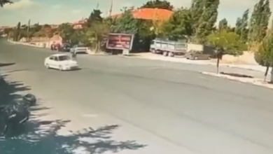 Görselin üst bölümünde, gündüz saatlerinde geniş bir caddeyi gören güvenlik kamerası kaydından bir an yer alıyor. Asfalt yol üzerinde seyir halinde olan beyaz renkli bir binek otomobil görülüyor. Yolun kenarında ve arka planda kiremit çatılı tek katlı yapılar, reklam panosu, park halindeki bir kamyonet ve ağaçlık alanlar seçilirken; sol üst köşede güvenlik kamerasına ait yeşil renkli tarih ve saat göstergesi yer alıyor.