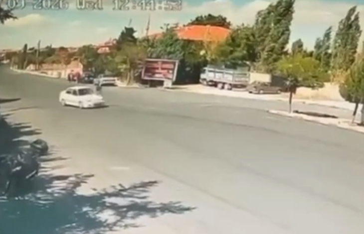 Görselin üst bölümünde, gündüz saatlerinde geniş bir caddeyi gören güvenlik kamerası kaydından bir an yer alıyor. Asfalt yol üzerinde seyir halinde olan beyaz renkli bir binek otomobil görülüyor. Yolun kenarında ve arka planda kiremit çatılı tek katlı yapılar, reklam panosu, park halindeki bir kamyonet ve ağaçlık alanlar seçilirken; sol üst köşede güvenlik kamerasına ait yeşil renkli tarih ve saat göstergesi yer alıyor.