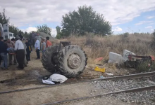 Görselin üst bölümünde, demiryolu hattı kenarında meydana gelen feci bir tren-traktör kazasının ardından oluşan enkaz ve olay yeri kalabalığı yer alıyor. Ön planda uzanan tren raylarının hemen yanında, çarpmanın şiddetiyle parçalanarak gövdesinden ayrılmış devasa traktör arka tekerlekleri ile sağ tarafa savrulmuş ve ters dönmüş traktör enkazı görülüyor. Sol tarafta ise beyaz bir minibüsün yanında toplanan köylüler, vatandaşlar ve yaralıya müdahale eden kalabalık bir grup duruyor. Çevrede kuru otlar, ağaçlar ve hafif bulutlu gökyüzü dikkat çekiyor.