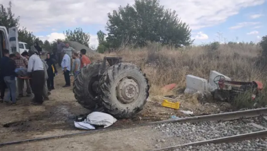 Görselin üst bölümünde, demiryolu hattı kenarında meydana gelen feci bir tren-traktör kazasının ardından oluşan enkaz ve olay yeri kalabalığı yer alıyor. Ön planda uzanan tren raylarının hemen yanında, çarpmanın şiddetiyle parçalanarak gövdesinden ayrılmış devasa traktör arka tekerlekleri ile sağ tarafa savrulmuş ve ters dönmüş traktör enkazı görülüyor. Sol tarafta ise beyaz bir minibüsün yanında toplanan köylüler, vatandaşlar ve yaralıya müdahale eden kalabalık bir grup duruyor. Çevrede kuru otlar, ağaçlar ve hafif bulutlu gökyüzü dikkat çekiyor.