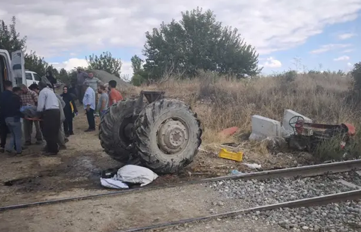Görselin üst bölümünde, demiryolu hattı kenarında meydana gelen feci bir tren-traktör kazasının ardından oluşan enkaz ve olay yeri kalabalığı yer alıyor. Ön planda uzanan tren raylarının hemen yanında, çarpmanın şiddetiyle parçalanarak gövdesinden ayrılmış devasa traktör arka tekerlekleri ile sağ tarafa savrulmuş ve ters dönmüş traktör enkazı görülüyor. Sol tarafta ise beyaz bir minibüsün yanında toplanan köylüler, vatandaşlar ve yaralıya müdahale eden kalabalık bir grup duruyor. Çevrede kuru otlar, ağaçlar ve hafif bulutlu gökyüzü dikkat çekiyor.