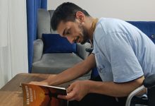 Yusuf Bingöl, ahşap bir masada oturmuş, dikkatle kitabını inceliyor. Üzerinde açık mavi bir tişört var. Elinde tuttuğu kitabın kapağında, kollarını gökyüzüne uzatan bir figür ve gün batımını andıran sıcak renkler yer alıyor. Kapakta “Bir Umudun Hikâyesi” başlığı görülüyor. Arkada gri bir koltuk, mavi minderler ve mavi bir perde bulunuyor. Ortam sade, sıcak ve ev içi bir çalışma alanı hissi veriyor.