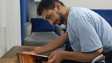 Yusuf Bingöl, ahşap bir masada oturmuş, dikkatle kitabını inceliyor. Üzerinde açık mavi bir tişört var. Elinde tuttuğu kitabın kapağında, kollarını gökyüzüne uzatan bir figür ve gün batımını andıran sıcak renkler yer alıyor. Kapakta “Bir Umudun Hikâyesi” başlığı görülüyor. Arkada gri bir koltuk, mavi minderler ve mavi bir perde bulunuyor. Ortam sade, sıcak ve ev içi bir çalışma alanı hissi veriyor.