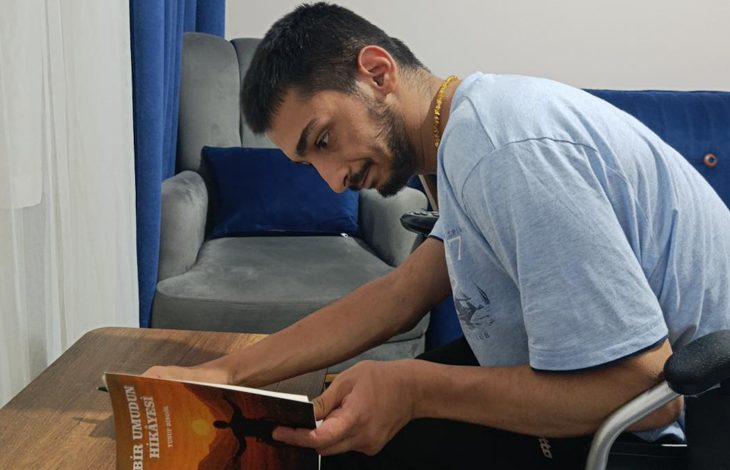 Yusuf Bingöl, ahşap bir masada oturmuş, dikkatle kitabını inceliyor. Üzerinde açık mavi bir tişört var. Elinde tuttuğu kitabın kapağında, kollarını gökyüzüne uzatan bir figür ve gün batımını andıran sıcak renkler yer alıyor. Kapakta “Bir Umudun Hikâyesi” başlığı görülüyor. Arkada gri bir koltuk, mavi minderler ve mavi bir perde bulunuyor. Ortam sade, sıcak ve ev içi bir çalışma alanı hissi veriyor.
