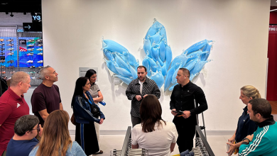 Mağaza içinde, adidas İstinyePark AVM’nin modern ve ferah tasarımında bir grup global ve Türkiye yöneticisi yarım daire şeklinde toplanmış durumda. Ön tarafta Mehmet Kızıltaş, Engelsiz Kariyer’in +1 Yetenek Engelli İş Koçu Programını konuklara anlatıyor. Arka planda duvara yerleştirilmiş adidas logosunu ifade eden büyük, mavi tonlarda yaprak formunda bir sanat enstalasyonu dikkat çekiyor. Çevrede raflarda sergilenen ürünler ve mağazanın ahşap detaylı mimarisi, profesyonel bir eğitim ve bilgilendirme buluşması atmosferi oluşturuyor.