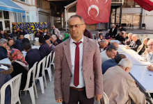 Görselin üst bölümünde, açık alanda kurulan uzun masalar etrafında toplanmış kalabalık bir topluluk ve ön planda ayakta duran Altı Nokta Körler Derneği Kütahya Şube Başkanı İsmail Barış yer alıyor. Görme engelli olan İsmail Barış; gözlükleri, açık kahverengi kırçıllı ceketi, beyaz gömleği ve bordo kravatıyla doğrudan öne doğru duruyor. Arkasında beyaz plastik sandalyelerde ve uzun ziyafet masalarında yan yana oturmuş kadın ve erkek dernek üyeleri ile misafirler sohbet ediyor. Arka fonda asılı büyük bir Türk bayrağı, sokak grafiti yazıları ve binalar görülüyor.