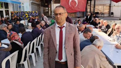 Görselin üst bölümünde, açık alanda kurulan uzun masalar etrafında toplanmış kalabalık bir topluluk ve ön planda ayakta duran Altı Nokta Körler Derneği Kütahya Şube Başkanı İsmail Barış yer alıyor. Görme engelli olan İsmail Barış; gözlükleri, açık kahverengi kırçıllı ceketi, beyaz gömleği ve bordo kravatıyla doğrudan öne doğru duruyor. Arkasında beyaz plastik sandalyelerde ve uzun ziyafet masalarında yan yana oturmuş kadın ve erkek dernek üyeleri ile misafirler sohbet ediyor. Arka fonda asılı büyük bir Türk bayrağı, sokak grafiti yazıları ve binalar görülüyor.