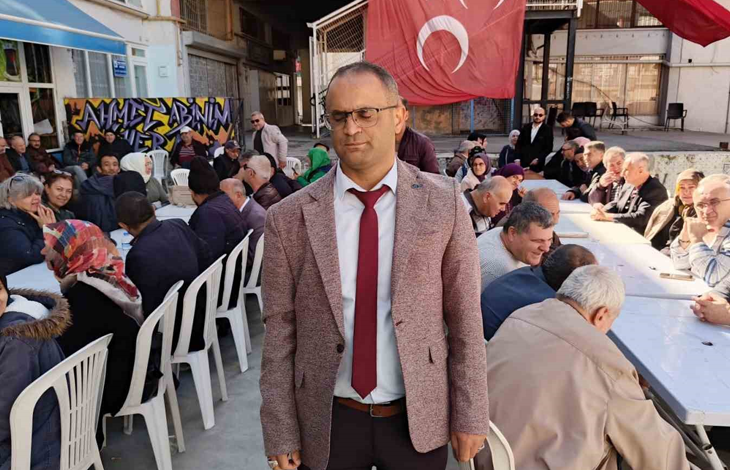 Görselin üst bölümünde, açık alanda kurulan uzun masalar etrafında toplanmış kalabalık bir topluluk ve ön planda ayakta duran Altı Nokta Körler Derneği Kütahya Şube Başkanı İsmail Barış yer alıyor. Görme engelli olan İsmail Barış; gözlükleri, açık kahverengi kırçıllı ceketi, beyaz gömleği ve bordo kravatıyla doğrudan öne doğru duruyor. Arkasında beyaz plastik sandalyelerde ve uzun ziyafet masalarında yan yana oturmuş kadın ve erkek dernek üyeleri ile misafirler sohbet ediyor. Arka fonda asılı büyük bir Türk bayrağı, sokak grafiti yazıları ve binalar görülüyor.