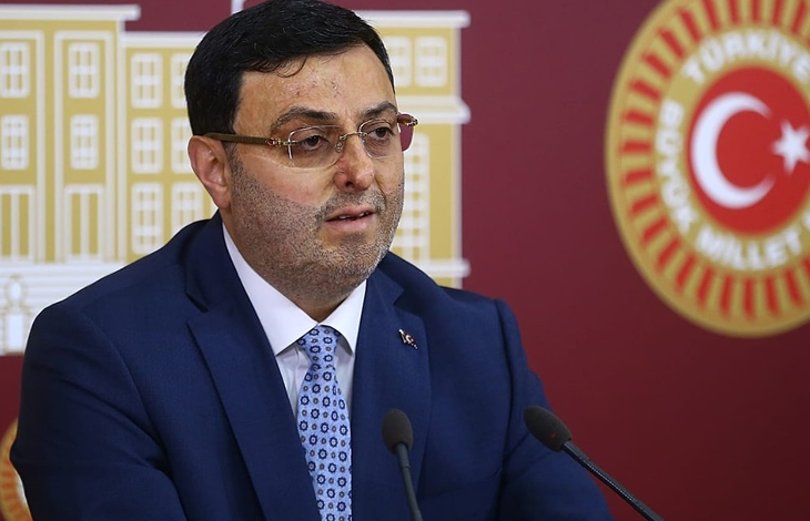 AK Parti İstanbul Milletvekili Serkan Bayram, TBMM’de düzenlenen bir basın açıklaması sırasında kürsüde konuşuyor. Üzerinde koyu renk takım elbise, beyaz gömlek ve desenli mavi bir kravat bulunuyor. Arka planda Türkiye Büyük Millet Meclisi amblemleri ve Türk bayrağı yer alıyor.
