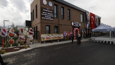 Görselde, önünde çelenkler ve çiçeklerle süslenmiş, Türk bayraklarının asılı olduğu modern bir kamu binasının açılış töreni için hazırlıklar yapıldığı görülmektedir.