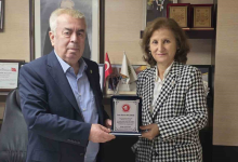 Görselin üst bölümünde, kurumsal bir çalışma ofisinde bir araya gelerek teşekkür plaketi takdim eden ve birlikte tutarak poz veren bir erkek ve bir kadın yer alıyor. Sol tarafta lacivert takım elbise ve açık mavi gömlek giymiş beyaz saçlı beyefendi ile sağ tarafta siyah-beyaz kazayağı desenli şık bir ceket ve beyaz bluz giymiş hanımefendi, ortalarında duran gümüş detaylı plaketi iki elleriyle tutarak objektife gülümsüyor. Arka plandaki ahşap ofis mobilyalarının üzerinde çerçeveli teşekkür belgeleri, ödüller ve masa üstü Türk bayrağı ile kurumsal flamalar görülüyor.