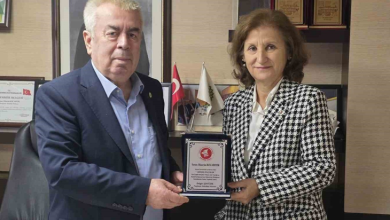 Görselin üst bölümünde, kurumsal bir çalışma ofisinde bir araya gelerek teşekkür plaketi takdim eden ve birlikte tutarak poz veren bir erkek ve bir kadın yer alıyor. Sol tarafta lacivert takım elbise ve açık mavi gömlek giymiş beyaz saçlı beyefendi ile sağ tarafta siyah-beyaz kazayağı desenli şık bir ceket ve beyaz bluz giymiş hanımefendi, ortalarında duran gümüş detaylı plaketi iki elleriyle tutarak objektife gülümsüyor. Arka plandaki ahşap ofis mobilyalarının üzerinde çerçeveli teşekkür belgeleri, ödüller ve masa üstü Türk bayrağı ile kurumsal flamalar görülüyor.