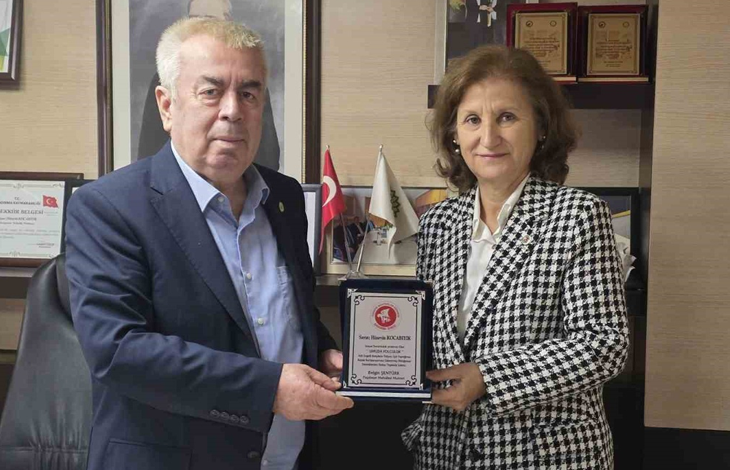 Görselin üst bölümünde, kurumsal bir çalışma ofisinde bir araya gelerek teşekkür plaketi takdim eden ve birlikte tutarak poz veren bir erkek ve bir kadın yer alıyor. Sol tarafta lacivert takım elbise ve açık mavi gömlek giymiş beyaz saçlı beyefendi ile sağ tarafta siyah-beyaz kazayağı desenli şık bir ceket ve beyaz bluz giymiş hanımefendi, ortalarında duran gümüş detaylı plaketi iki elleriyle tutarak objektife gülümsüyor. Arka plandaki ahşap ofis mobilyalarının üzerinde çerçeveli teşekkür belgeleri, ödüller ve masa üstü Türk bayrağı ile kurumsal flamalar görülüyor.