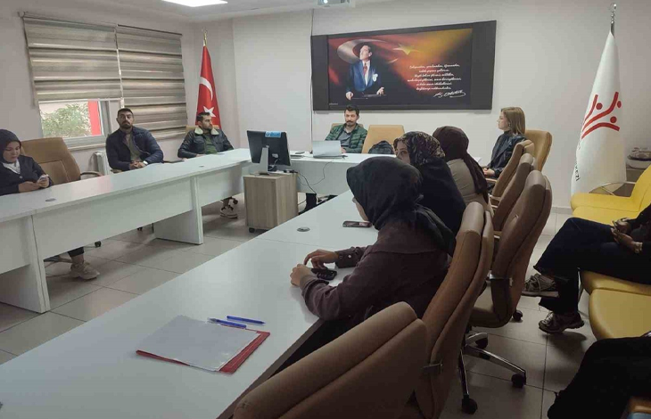 Instagram İçin Alternatif Metin: Görselin üst bölümünde, kurumsal bir toplantı odasında U şeklinde dizilmiş beyaz masaların etrafında oturan Engelli ve Yaşlı Hizmetleri personeli yer alıyor. Masalarda not dosyaları, kalemler ve bilgisayarlar bulunurken; başörtülü ve takım kıyafetli kadın ile erkek personeller dikkatle toplantıyı takip ediyor. Odanın baş tarafındaki duvarda Atatürk portresi ve vecizesi asılı duruyor; köşede Türk bayrağı ile sağ tarafta Aile ve Sosyal Hizmetler Bakanlığı'nın logosunu taşıyan beyaz bayrak görülüyor.