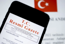 Görselde, yakın plan çekilmiş bir akıllı telefon ekranında T.C. Resmî Gazete ibaresi ve yayım detayları yer almaktadır; arka planda ise Türk bayrağı silüeti ve yazılı kağıtlar bulunmaktadır.