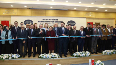 Görselde, geniş bir salon veya etkinlik alanında açılış kurdelesi kesmek üzere yan yana dizilmiş protokol üyeleri ve kalabalık bir katılımcı grubu yer almaktadır. Arkadaki afişte "ATLAS AZ GÖRME REHABİLİTASYON MERKEZİ" yazısı dikkat çekmektedir.