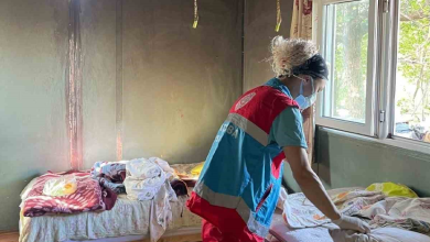 Görselde, mütevazı bir ev ortamında kırmızı ve mavi renkli resmi bakım veya yardım görevlisi yeleği giymiş, maskeli ve saçları dağınık bir kadın görevli, yatakları düzenlerken veya bakım çalışması yaparken görülmektedir. Oda içerisinde sol tarafta yatak ve örtüler yer alırken, sağ tarafta dışarıya bakan aydınlık bir pencere bulunmaktadır.