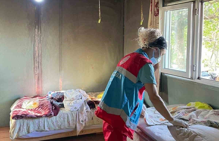 Görselde, mütevazı bir ev ortamında kırmızı ve mavi renkli resmi bakım veya yardım görevlisi yeleği giymiş, maskeli ve saçları dağınık bir kadın görevli, yatakları düzenlerken veya bakım çalışması yaparken görülmektedir. Oda içerisinde sol tarafta yatak ve örtüler yer alırken, sağ tarafta dışarıya bakan aydınlık bir pencere bulunmaktadır.