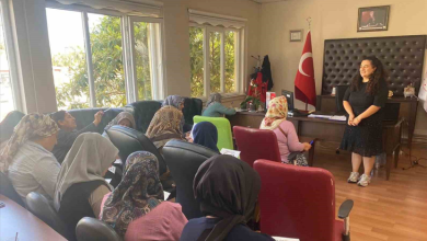 Görselde, aydınlık bir odada koltuklarda oturan katılımcı kadınlara sunum veya eğitim veren bir eğitmen yer almaktadır. Odanın köşesinde Türk bayrağı ve duvarda çerçeveli bir tablo bulunmaktadır.