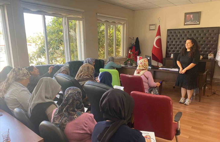 Görselde, aydınlık bir odada koltuklarda oturan katılımcı kadınlara sunum veya eğitim veren bir eğitmen yer almaktadır. Odanın köşesinde Türk bayrağı ve duvarda çerçeveli bir tablo bulunmaktadır.