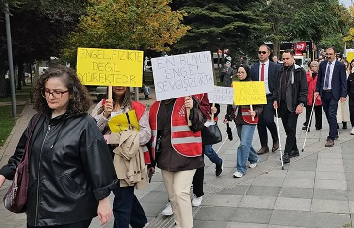 Görselin üst bölümünde, bir cadde üzerinde yürüyüş düzenleyen kalabalık bir grup yer almaktadır. Gruptakiler ellerinde "Engel Fizikte Değil Yürektedir" ve "En Büyük Engel Sevgisizliktir" yazılı pankartlar taşırken, bazı görme engelli katılımcılar beyaz bastonlarıyla yürüyüşe eşlik etmektedir.