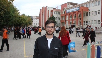 Görselin üst bölümünde, bir okulun bahçesinde, arkasında okul binası ve öğrenciler bulunan görme engelli genç bir öğretmen yer almaktadır. Siyah fermuarlı bir spor ceket giyen ve gözlük takan öğretmen, doğrudan kameraya bakarak hafifçe gülümsemektedir. Arka plandaki okul binasının üzerinde "Fatih Sultan Mehmet Ortaokulu" tabelası okunurken; bahçede okul formalı öğrenciler dolaşmakta, öğretmenler ve hareketli bir okul ortamı göze çarpmaktadır.
