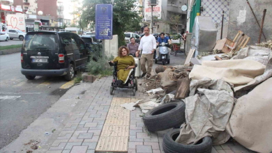 Görselin üst bölümünde, şehir merkezindeki bir kaldırımda akülü tekerlekli sandalyesiyle ilerlemeye çalışan ortopedik engelli bir kadın ve çevresindeki engeller yer alıyor. Kadının üzerinde hardal sarısı bir takım bulunurken, tekerlekli sandalyesiyle kaldırım ortasındaki sarı renkli hissedilebilir görme engelli kılavuz izlerinin üzerinde duruyor. Kaldırımın sağ tarafında yığılmış eski araba lastikleri, çuvallar, inşaat atıkları ve brandalar yürüyüş yolunu daraltırken; arka planda yürüyen yayalar, motosikletli bir sürücü, park halindeki siyah bir hafif ticari araç ve cadde üzerindeki binalar görülüyor.