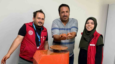 Instagram İçin Alternatif Metin: Görselin üst bölümünde, çömlek atölyesinde elektrikli çömlek çarkı makinesinin arkasında yan yana durarak poz veren üç kişi yer alıyor. Ortada çizgili polo yaka tişört giymiş engelli birey, iki avucunun içinde kilden yaptığı minik çömlek kabını gururla ileriye uzatarak sergiliyor. Solunda kırmızı Gençlik ve Spor Bakanlığı yeleği giymiş, elleri kile bulanmış eğitmen genç adam; sağında ise koyu yeşil başörtülü ve aynı kırmızı yelekten giymiş gülümseyen genç bir kadın görevli duruyor. Önlerinde turuncu gövdeli döner çömlek çarkı ve açık renkli sade duvar arka planı görülüyor.