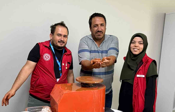 Instagram İçin Alternatif Metin: Görselin üst bölümünde, çömlek atölyesinde elektrikli çömlek çarkı makinesinin arkasında yan yana durarak poz veren üç kişi yer alıyor. Ortada çizgili polo yaka tişört giymiş engelli birey, iki avucunun içinde kilden yaptığı minik çömlek kabını gururla ileriye uzatarak sergiliyor. Solunda kırmızı Gençlik ve Spor Bakanlığı yeleği giymiş, elleri kile bulanmış eğitmen genç adam; sağında ise koyu yeşil başörtülü ve aynı kırmızı yelekten giymiş gülümseyen genç bir kadın görevli duruyor. Önlerinde turuncu gövdeli döner çömlek çarkı ve açık renkli sade duvar arka planı görülüyor.