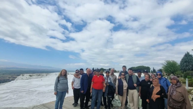 Görselin üst bölümünde, Denizli’nin ünlü beyaz travertenleri önünde gezi amaçlı bir arada durarak gülümseyen engelli bireyler, aileleri ve rehberlerden oluşan kalabalık bir grup yer alıyor. Açık havada, taş döşeli teras alanı üzerinde duran katılımcıların arkasında panoramik ova manzarası ve bulutlu mavi gökyüzü uzanıyor.