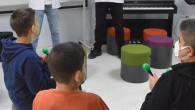 Görselde, kapalı bir odada ellerinde küçük renkli enstrümanlar (marakas) tutarak oturan ve müzik etkinliğine katılan çocuklar yer almaktadır; arka planda ise ayakta duran eğitmenler ve piyanolar bulunmaktadır.