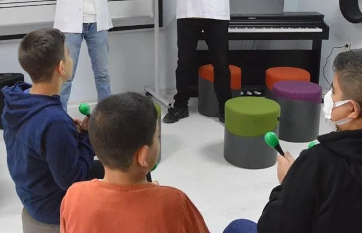 Görselde, kapalı bir odada ellerinde küçük renkli enstrümanlar (marakas) tutarak oturan ve müzik etkinliğine katılan çocuklar yer almaktadır; arka planda ise ayakta duran eğitmenler ve piyanolar bulunmaktadır.