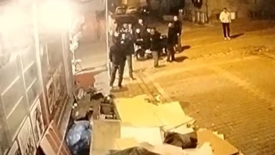 Görselde, gece vakti sokakta güvenlik kamerası açısıyla yukarıdan çekilmiş bir görüntü yer almaktadır. Bir binanın önünde veya dükkan kenarında toplanmış bir grup insan ve etrafta karton parçaları, çöpler görülmektedir.