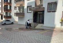 Görselde, apartman girişinin önünde merdivenli bir bina girişi, kaldırımda elektrikli tekerlekli sandalye kullanan Furkan Ulusoy, çevredeki çok katlı apartmanlar görülüyor.