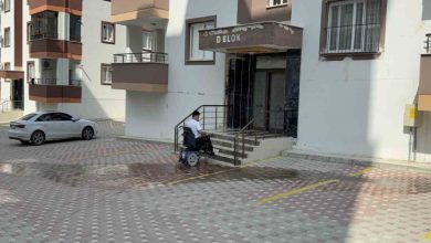 Görselde, apartman girişinin önünde merdivenli bir bina girişi, kaldırımda elektrikli tekerlekli sandalye kullanan Furkan Ulusoy, çevredeki çok katlı apartmanlar görülüyor.