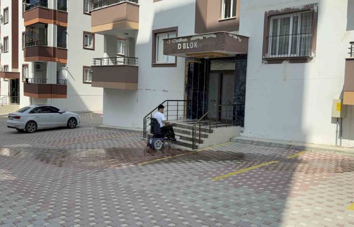 Görselde, apartman girişinin önünde merdivenli bir bina girişi, kaldırımda elektrikli tekerlekli sandalye kullanan Furkan Ulusoy, çevredeki çok katlı apartmanlar görülüyor.