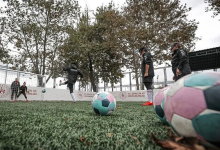 Görselin üst bölümünde, açık havadaki suni çim sahada antrenman yapan Görme Engelli Kadın Milli Futbol Takımı oyuncuları yer alıyor. Ön planda çim zemin üzerinde duran pembe, mavi ve beyaz desenli çıngıraklı futbol topları odak noktası oluştururken; arka planda göz bantları takılı milli sporcular topa vuruş ve pas çalışması yapıyor. Sahayı çevreleyen bordürlerde T.C. Gençlik ve Spor Bakanlığı logoları, kalenin önünde hazır bekleyen kaleci ile arkada sonbahar renklerine bürünmüş ağaçlar görülüyor.