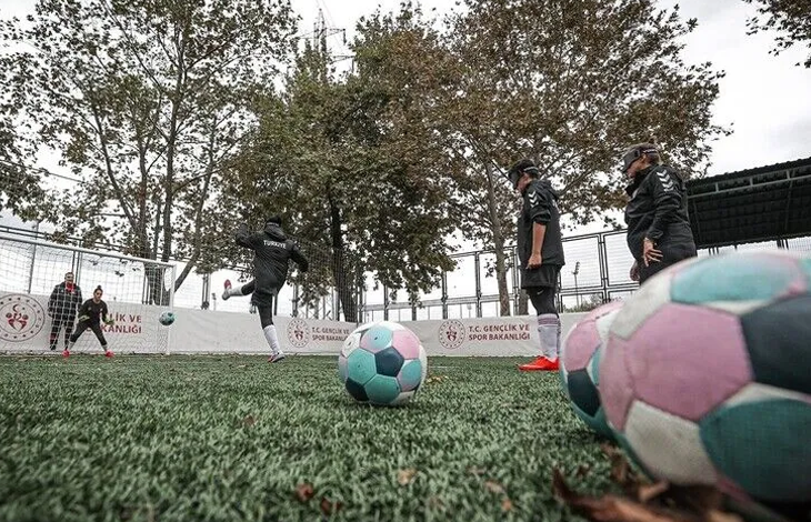 Görselin üst bölümünde, açık havadaki suni çim sahada antrenman yapan Görme Engelli Kadın Milli Futbol Takımı oyuncuları yer alıyor. Ön planda çim zemin üzerinde duran pembe, mavi ve beyaz desenli çıngıraklı futbol topları odak noktası oluştururken; arka planda göz bantları takılı milli sporcular topa vuruş ve pas çalışması yapıyor. Sahayı çevreleyen bordürlerde T.C. Gençlik ve Spor Bakanlığı logoları, kalenin önünde hazır bekleyen kaleci ile arkada sonbahar renklerine bürünmüş ağaçlar görülüyor.
