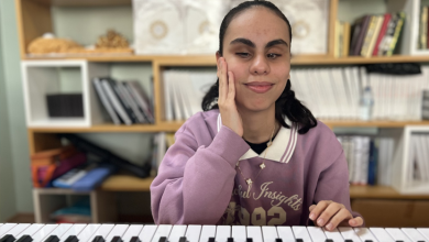 Instagram İçin Alternatif Metin: Görselin üst bölümünde, bir müzik odasında klavyenin (synthesizer) başında oturarak hafifçe gülümseyen görme engelli müzisyen Duygu Murcak yer alıyor. Üzerinde lila renkli kapüşonlu veya sweatshirt tarzı bir üst giysi bulunan genç kadın, sağ elini yanağına dayamış vaziyette poz verirken sol eliyle klavyenin tuşlarına dokunuyor. Arkasındaki ahşap raflı kitaplıkta düzenli bir şekilde dizilmiş dosyalar, kitaplar ve çeşitli eşyalar dikkat çekiyor.
