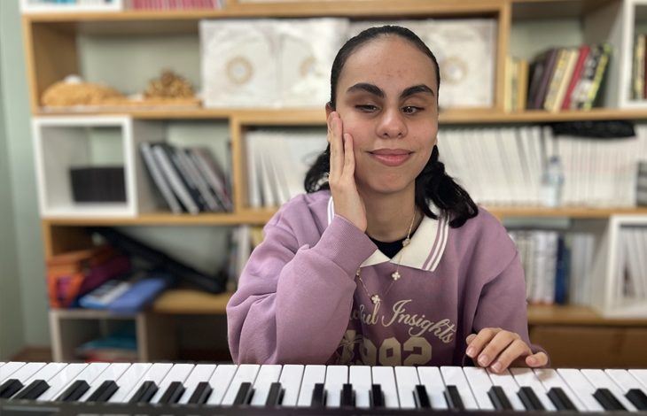 Instagram İçin Alternatif Metin: Görselin üst bölümünde, bir müzik odasında klavyenin (synthesizer) başında oturarak hafifçe gülümseyen görme engelli müzisyen Duygu Murcak yer alıyor. Üzerinde lila renkli kapüşonlu veya sweatshirt tarzı bir üst giysi bulunan genç kadın, sağ elini yanağına dayamış vaziyette poz verirken sol eliyle klavyenin tuşlarına dokunuyor. Arkasındaki ahşap raflı kitaplıkta düzenli bir şekilde dizilmiş dosyalar, kitaplar ve çeşitli eşyalar dikkat çekiyor.
