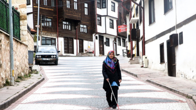 Görselin üst bölümünde, tarihi konakların ve geleneksel evlerin bulunduğu, Arnavut kaldırımlı yokuş bir sokakta beyaz uzun bastonuyla yürüyen görme engelli Nezihe Dalahmetoğlu yer almaktadır. Siyah kıyafetler giymiş ve renkli bir başörtüsü takmış olan Dalahmetoğlu, bastonunu kullanarak güvenli bir şekilde yürürken sokak dokusu ve park etmiş bir araç arka planda dikkat çekmektedir.