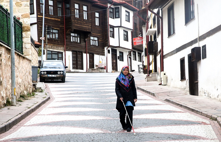 Görselin üst bölümünde, tarihi konakların ve geleneksel evlerin bulunduğu, Arnavut kaldırımlı yokuş bir sokakta beyaz uzun bastonuyla yürüyen görme engelli Nezihe Dalahmetoğlu yer almaktadır. Siyah kıyafetler giymiş ve renkli bir başörtüsü takmış olan Dalahmetoğlu, bastonunu kullanarak güvenli bir şekilde yürürken sokak dokusu ve park etmiş bir araç arka planda dikkat çekmektedir.