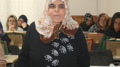 Görselde, kapalı bir alanda ön planda yer alan, desenli başörtülü ve koyu renk kıyafetli bir kadın ile arka planda masalarda oturarak ders çalışan veya kitap okuyan diğer katılımcılar yer almaktadır.