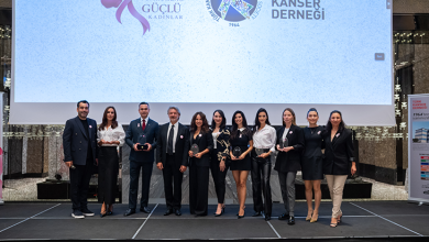 Sahnede yan yana dizilmiş on bir kişi bulunuyor. Hepsi resmî ve şık kıyafetler giymiş; bazıları ellerinde cam plaketler veya ödüller tutuyor. Arkalarında büyük bir ekran yer alıyor. Ekranda pembe bir kurdele grafiği ve şu ifadeler görünüyor: “Güçlü Yumruklar Güçlü Kadınlar” “Türk Kanser Derneği / Turkish Cancer Society” “1964’ten beri yanınızda...” Sahne profesyonel bir etkinlik alanını andırıyor; ışıklandırma ve dekor düzenli. Genel atmosfer, kanser farkındalığı ve kadınlara destek temalı bir ödül veya tanıtım etkinliği izlenimi veriyor.