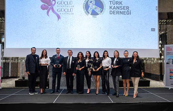 Sahnede yan yana dizilmiş on bir kişi bulunuyor. Hepsi resmî ve şık kıyafetler giymiş; bazıları ellerinde cam plaketler veya ödüller tutuyor. Arkalarında büyük bir ekran yer alıyor. Ekranda pembe bir kurdele grafiği ve şu ifadeler görünüyor: “Güçlü Yumruklar Güçlü Kadınlar” “Türk Kanser Derneği / Turkish Cancer Society” “1964’ten beri yanınızda...” Sahne profesyonel bir etkinlik alanını andırıyor; ışıklandırma ve dekor düzenli. Genel atmosfer, kanser farkındalığı ve kadınlara destek temalı bir ödül veya tanıtım etkinliği izlenimi veriyor.