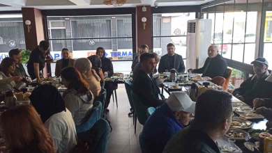 Görselde, aydınlık bir restoranda uzun bir masa etrafında toplanmış, kahvaltı yapan kalabalık bir grup yer almaktadır. Katılımcılar masada sohbet ederek kahvaltı yaparken, geniş pencerelerden dışarıdaki cadde görünmektedir.