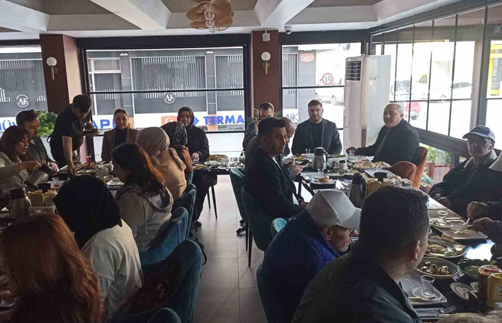 Görselde, aydınlık bir restoranda uzun bir masa etrafında toplanmış, kahvaltı yapan kalabalık bir grup yer almaktadır. Katılımcılar masada sohbet ederek kahvaltı yaparken, geniş pencerelerden dışarıdaki cadde görünmektedir.