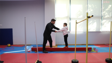 Görselde, kapalı spor salonunda bir eğitmen ile çocuk yer almaktadır. Eğitmen, denge tahtası üzerinde duran çocuğun ellerinden tutarak ona destek olmaktadır.